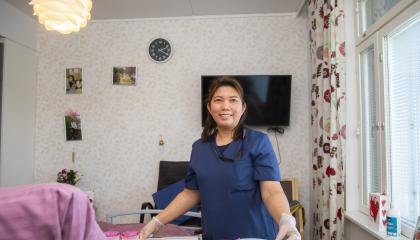 Filippiiniläinen hoiva-avustaja seisoo hymyillen asukkaan huoneessa.