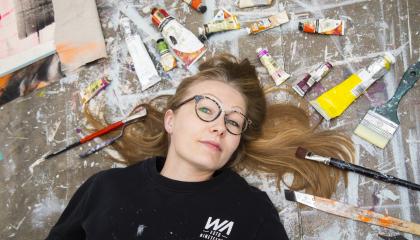 Silmälasipäinen nainen makaa lattialla. Hänen hiuksensa ovat pitkin lattiaa. Hiuksia kehystää lattialla olevat maalituubit ja pensselit. Lattialla on maaliroiskeita.