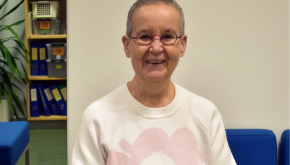 Henkilö istuu sohvalla ja katsoo kameraan. Taustalla peili, jossa näkyy mappeja.