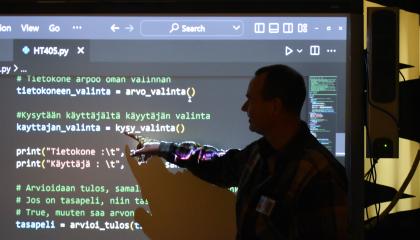 Henkilö seisoo älytaulun edessä. Taululla on Python-kielistä ohjelmointitekstiä.