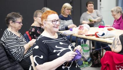 Kahdeksan ihmistä istuu salissa neulomassa käsitöitä