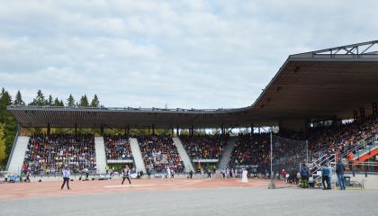 Pesäpallokentällä peli käynnissä etualalla ja takana näkyy katsomo, jossa väkeä istumassa.