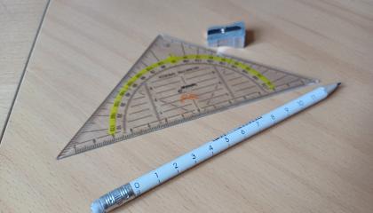 Kuvassa kynä, teroitin ja piirtokolmio.