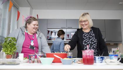 Kaksi henkilöä seisoo tarjoiluastioita täynnä olevan buffetpöydän takana.