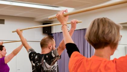 Kolme henkilöä keppijumppaa liikuntasalissa. Ylös nostetut kädet pitelevät puista keppiä.