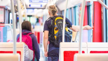 Kaksi henkilöä seisoo ratikkavaunun käytävällä selin. Etualalla on istuimia ja taustalla reittitaulu.