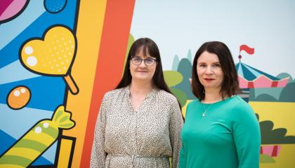 Tampereen kaupungin lapsiasiamiehet Tiia Heinäsuo ja Taru Kuosmanen seisovat rinnakkain. Heidän takanaan on värikäs, voimakkaasti kuvioitu seinä, jossa erottuu karkkeja ja sirkusteltta.