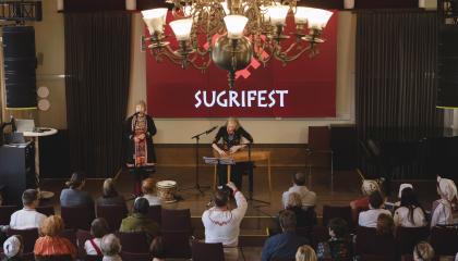 Suuren juhlasalin lavalla esiintyy laulaja ja kanteleen soittaja. Yleisö istuu penkeillä.