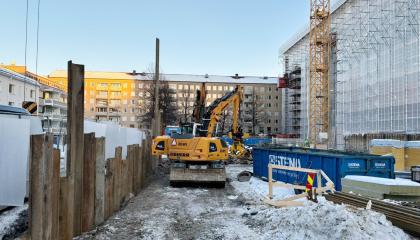 Virastotalon takapihalla tehdään työkoneen avulla rakentamistöitä tukiseinän ja suojahuppuisen talon välissä.