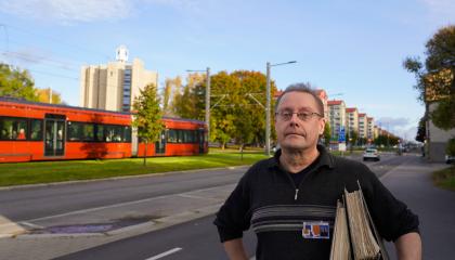 Henkilö seisoo kadulla. Hänen kainalossaan on arkistomappeja. Ratikka kulkee kadulla taustalla.