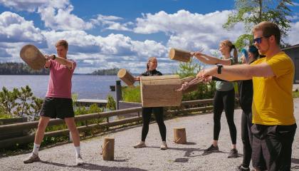 Viisi ihmistä kuntoilee köyden päässä olevia pöllejä heilutellen kesäisen järven rannalla.