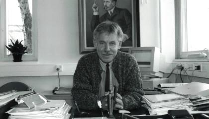 Henkilö istuu työhuoneessaan. Taustalla on suuri taulu ja pöydällä runsaasti papereita.