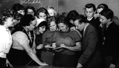 Joukko opiskelijoita on kokoontunut opettajan ympärille tutkimaan kirjaa.