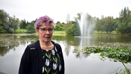 Sirkka-Liisa Jukarainen seisoo puistossa. Takana on lampi ja suihkulähde.