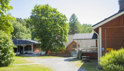 Lehtipuiden ympäröimä piha, jonka ympärillä rakennuksia. Oikealla punaisen hirsitalon pääty, keskellä punainen harjakattoinen rakennus. Vasemmalla matala punainen rakennus, jonka edessä aidattu terassi. Rakennuksessa avokuisti. Pihalla rakennusten edessä pöytiä ja tuoleja.