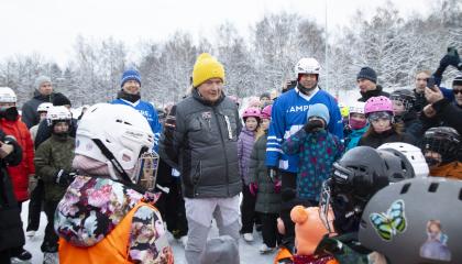 Iso joukko ihmisiä jäällä, suurin osa lapsia. Keskellä aikuinen mies keltainen pipo päässä, hänen molemmilla puolillaan seisoo mies. Useimmilla ihmisillä on päässään kypärä, joillakin pipo ja monilla on luistimet jalassa.