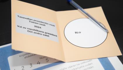 Tasolla ruskea äänestyslippu, jonka päällä kynä. Alla paperi, jossa numeromalleja.