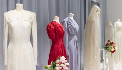 Hääpukuja mallinukkejen päällä näyttelytilassa. Puvut ovat pääasiassa valkoisia, mutta keskellä on sininen ja punainen hääpuku. Pukujen joukossa on kukkakimppuja.