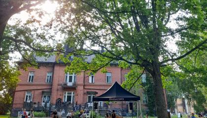 Kesäiseen puistoon on kokoontunut ihmisiä kuuntelemaan konserttia. Esiintyjä soittaa kosketinsoitinta telttakatoksen alla.