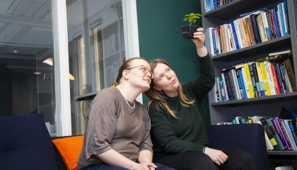 Kaksi naista istuu sohvalla. Oikeanpuoleinen pitää korkealla kädessään pientä ruukussa olevaa kasvia, jota molemmat katselevat. Oikealla puolella on kirjahylly.