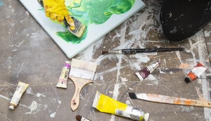 Lattialla maalituubeja, siveltimiä ja tekeillä oleva maalaus. Hanskaan verhottu käsi pitele sivellintä taulun päällä.