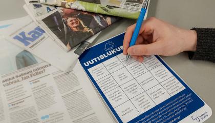 Uutislukubingon bingolomake pöydällä, jolla on myös sanomalehtiä. Lomakkeen yllä on käsi, joka pitelee kynää. 