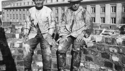 Kaksi hattupäistä muuraria istuu piipputiilien päällä.