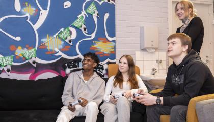 Neljä ihmistä huoneessa. Kolme istuu, yksi seisoo. Istuvilla on käsissään jokin laite. 