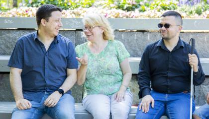 Kolme henkeä istuu rinnakkain puistossa keskustelemassa. Yhdellä heistä on näkövammaisen keppi ja mustat lasit.