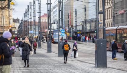 Katunäkymä. Rakennuksia, ihmisiä, raitiovaunu ja metallipylväitä.