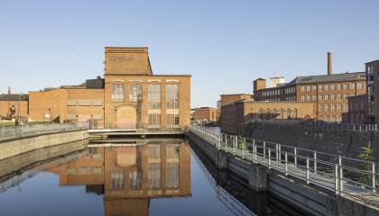 Museokeskus Vapriikki, punatiilinen kaksikerroksinen rakennus, kuvattuna ulkoa kevätiltana. Edessä patoaltaassa vettä, sivussa Tammerkoski. Taivas on pilvetön. 