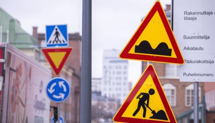 Liikennemerkkejä vasemmassa ja oikeassa laidassa. Taustalla kerrostaloja ja katu.