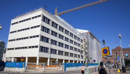 Valkoinen suuri rakennus on rakennustyömaa-aidan takana ja osittain vielä työmaahupun peitossa. Etualalla kävelee ihmisiä.