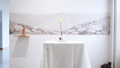 Valkoisen pöytäliinan päälle on asetetty oksannäköinen kynttilä. Seinälle on ripustettu piirustus kivikosta.