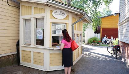 Amurin museokorttelin sisäänkäynnillä yksi henkilö ostamassa lippua luukulta. Taustalla museokorttelin rakennuksia ja ihmisiä istuu kahvilan pöydässä. Kesäinen, aurinkoinen ilma.