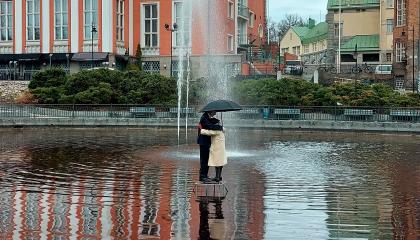 Valokuva kaupunkitilassa olevasta lammesta, jonka keskellä on vanhempaa pariskuntaa esittävä veistos. Pari seisoo sateenvarjon alla.