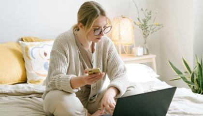 Nuori naishenkilö istuu sängyllä tietokoneen äärellä ja hänellä on kädessään puhelin.