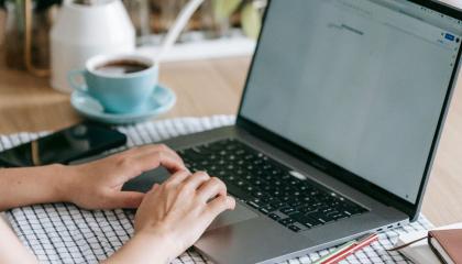 Pöydällä on kahvikuppi ja tietokone. Kädet ovat näppäimistöllä.