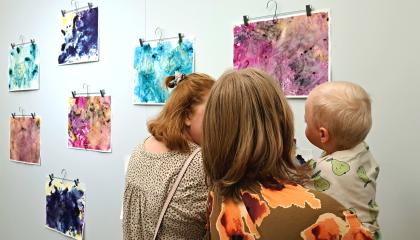 Seinällä abstrakteja vauvojen ja taaperoiden tekemiä taideteoksia. Kaksi aikuista ja toisella vauva sylissään ihastelemassa teoksia. 