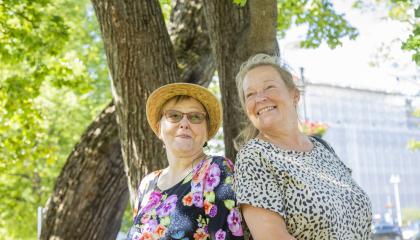 Kaksi hymyilevää naista nojaa puunrunkoon.