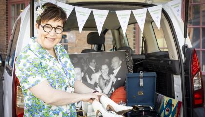 Tummahiuksinen, silmälasipäinen nainen näyttää valkoisia naisten korkokenkiä auton tavaratilan edessä.