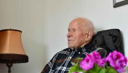 Henkilö istuu nojatuolissa. Hänen edessään on kukkakimppu ja vieressä pöytälamppu.