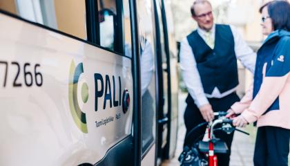 Palveluliikenteen avustaja auttaa rollaattoria käyttävää siirtymään Pali-bussin kyytiin.