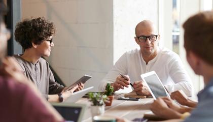 Neljä henkilöä keskustelevat kokouspöydän äärellä. Pöydällä on muistiinpanovälineitä kuten kannettavia tietokoneita, vihkoja ja kyniä. 