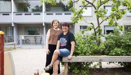 Kaksi työntekijää on kerrostalossa sijaitsevan päiväkodin pihalla. Edessä on leikkivälineitä ja takana kerrostalo.