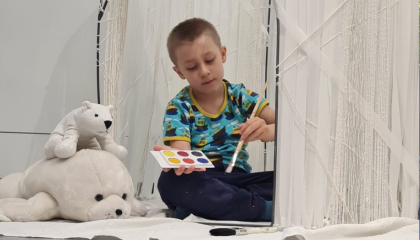 Lapsi istuu lattialla valkoisessa majassa ja maalaa majan valkoisista langoista tehtyä seinää eri värisillä vesiväreillä.