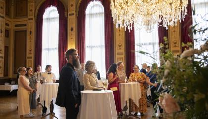 Ihmisiä suuressa salissa seisomassa pienten pyöreiden pöytien ääressä.