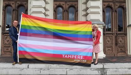Kaksi henkilöä seisoo Tampereen Raatihuoneen rappusilla. He levittävät näkyviin suurta Tampereen omaa pride-lippua, jossa on 13 väriraitaa.