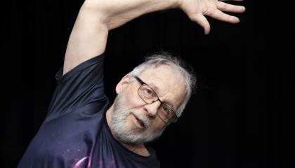 Mies mustaa taustaa vasten, vasen käsi ylhäällä taivutettuna pään päälle.