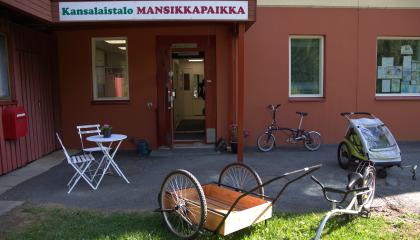 Erilaisia pyöriä ja maitokärryt Kansalaistalo Mansikkapaikan aurinkoisella etupihalla.